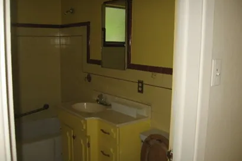 Tiny bathroom with a yellow vanity under a mirror, sink in front, and a tub/shower to the left. Window light in the back, wall switch nearby.