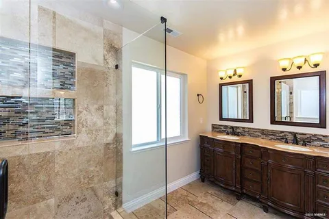 Bathroom with glass-enclosed walk-in shower on left, beige tile walls and floor, and a dark wood double vanity with three mirrors and warm light fixtures.