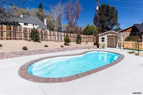 Kidney-shaped in-ground pool in a sunny backyard with brick coping, concrete decking, a small shed, and a wooden fence behind.