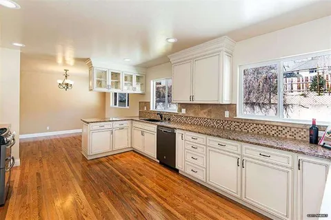 Bright kitchen with white cabinets, granite countertops, stainless steel dishwasher, and large windows along the counter; warm hardwood floors.