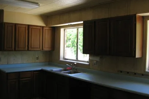 Dim kitchen with dark wood cabinets, pale countertops, and a double sink under a window; greenery visible outside.