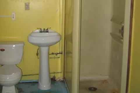 Small bathroom with a white toilet and pedestal sink, yellow walls, and a glass shower stall to the right; exposed pipes under the sink.