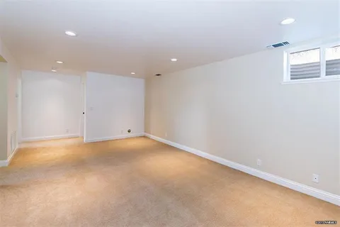Empty beige-carpeted basement with white walls, recessed ceiling lights, and a small high window at the far end.