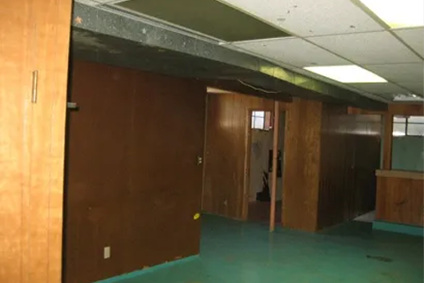 Empty office hallway with brown wood paneling, green carpet, and a drop ceiling; open doors along the corridor.