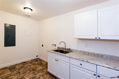A small white kitchen or utility area with granite counter, sink, white cabinets, and brown tiled floor, plus an electrical panel on the left wall and bright ceiling light