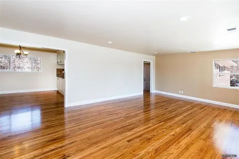 Empty living room with polished hardwood floors, light beige walls, white baseboards, and recessed lighting across the ceiling.