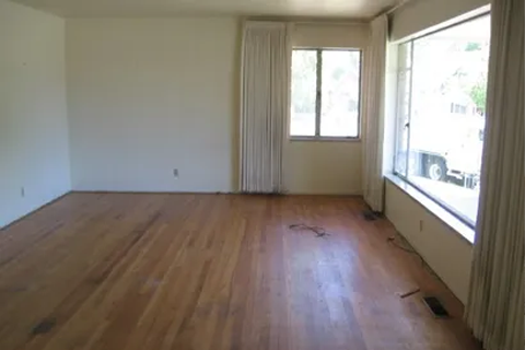 Empty living room with hardwood floors, light walls, and large windows with curtains letting in natural light.